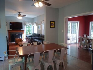 A spacious living room with a wooden dining table, white chairs, a fireplace, and a television.