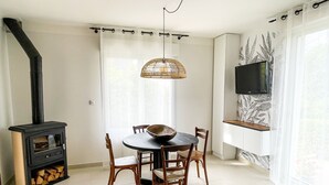 A dining area with a round table, four wooden chairs, a woven light fixture, a black wood-burning stove, and a TV mounted on the wall. - HOUSE GARDEN BEACHES WALKS (Saint-Nazaire)
