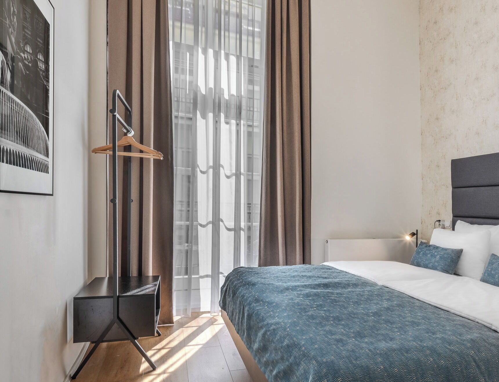 A modern bedroom with a bed, a bedside table, a hanging coat rack, and a window with curtains.