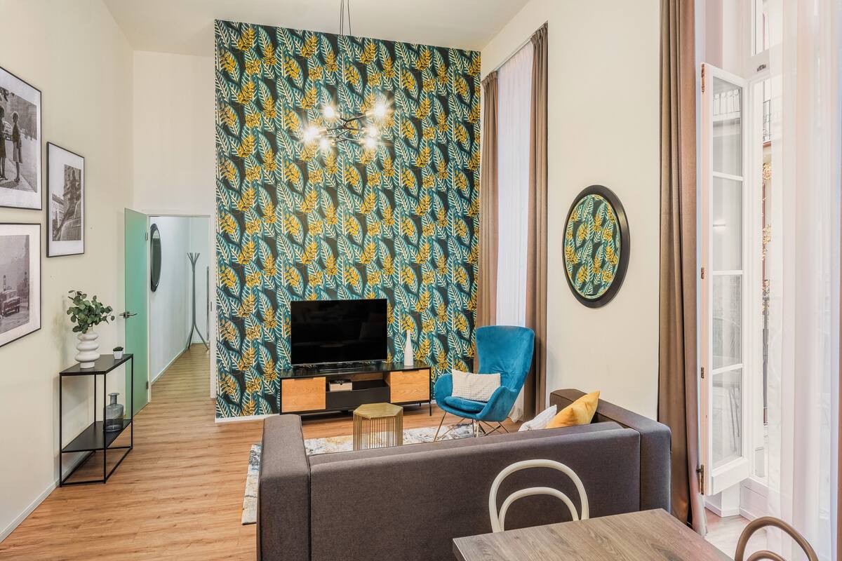 Uma sala de estar com sofá, televisão e uma poltrona azul. Há uma parede estampada atrás da TV e uma porta que dá para outro cômodo.
