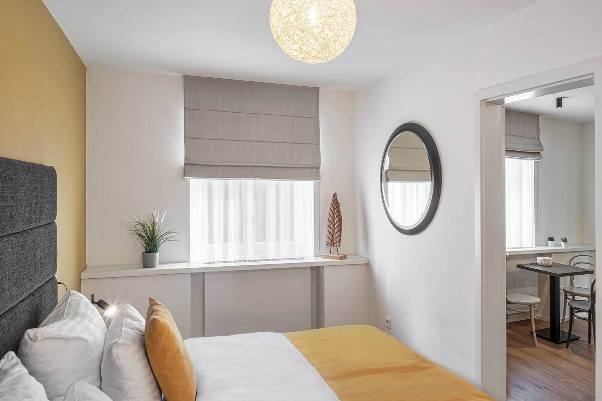 Quarto com cama, mesa de cabeceira, janela com cortinas e espelho na parede.