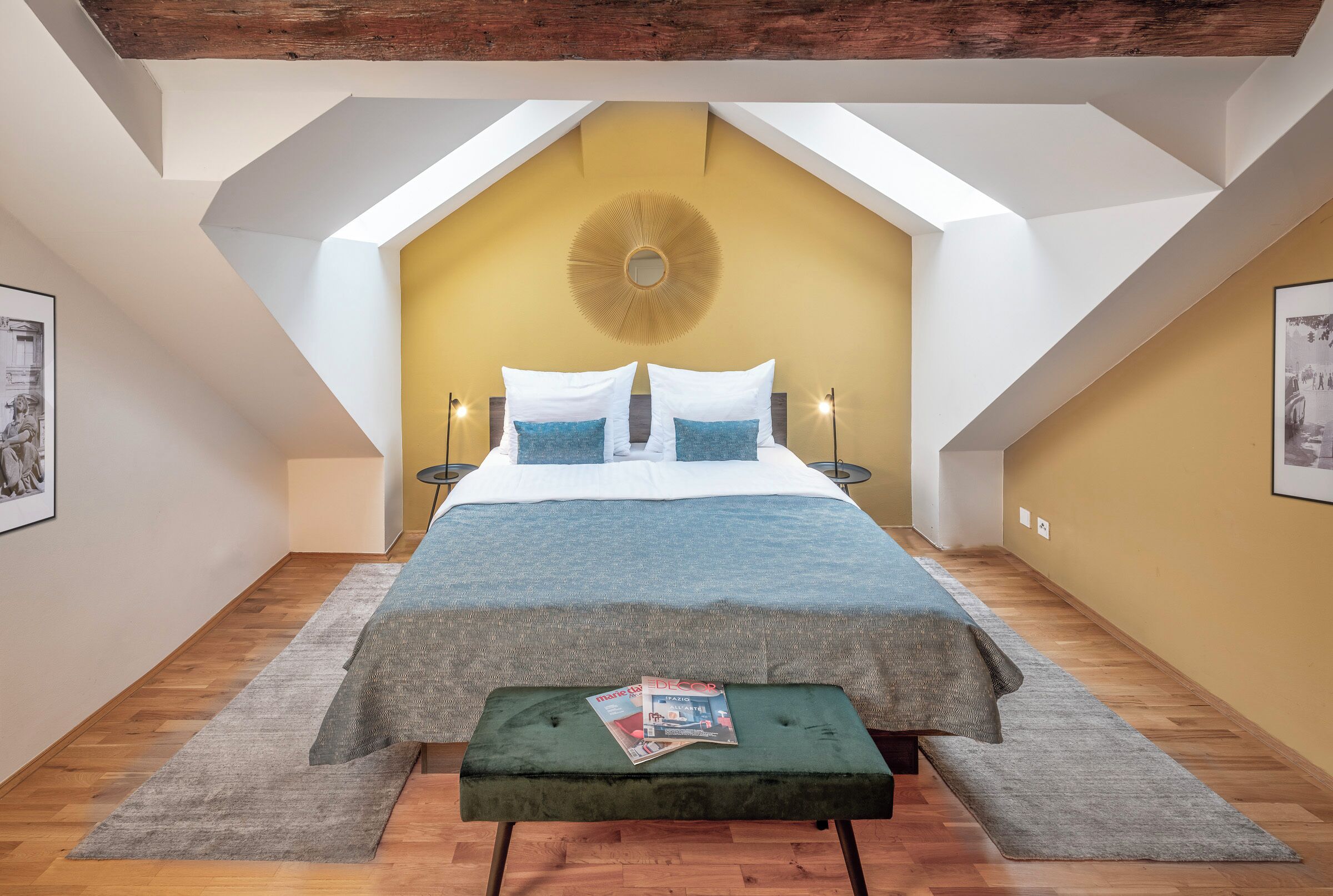 A bedroom with a sloped ceiling, a bed with white and blue bedding, a green bench, and two bedside tables with lamps.