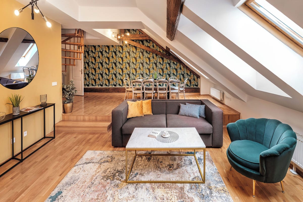 Um apartamento loft moderno com teto alto, vigas de madeira aparentes, escada, área de jantar com cadeiras, sofá e uma poltrona verde.