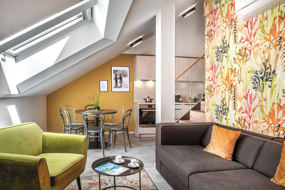 Uma sala de estar moderna com uma poltrona verde, um sofá cinza e uma mesa de centro. Há uma área de jantar com mesa e cadeiras, e uma cozinha ao fundo.