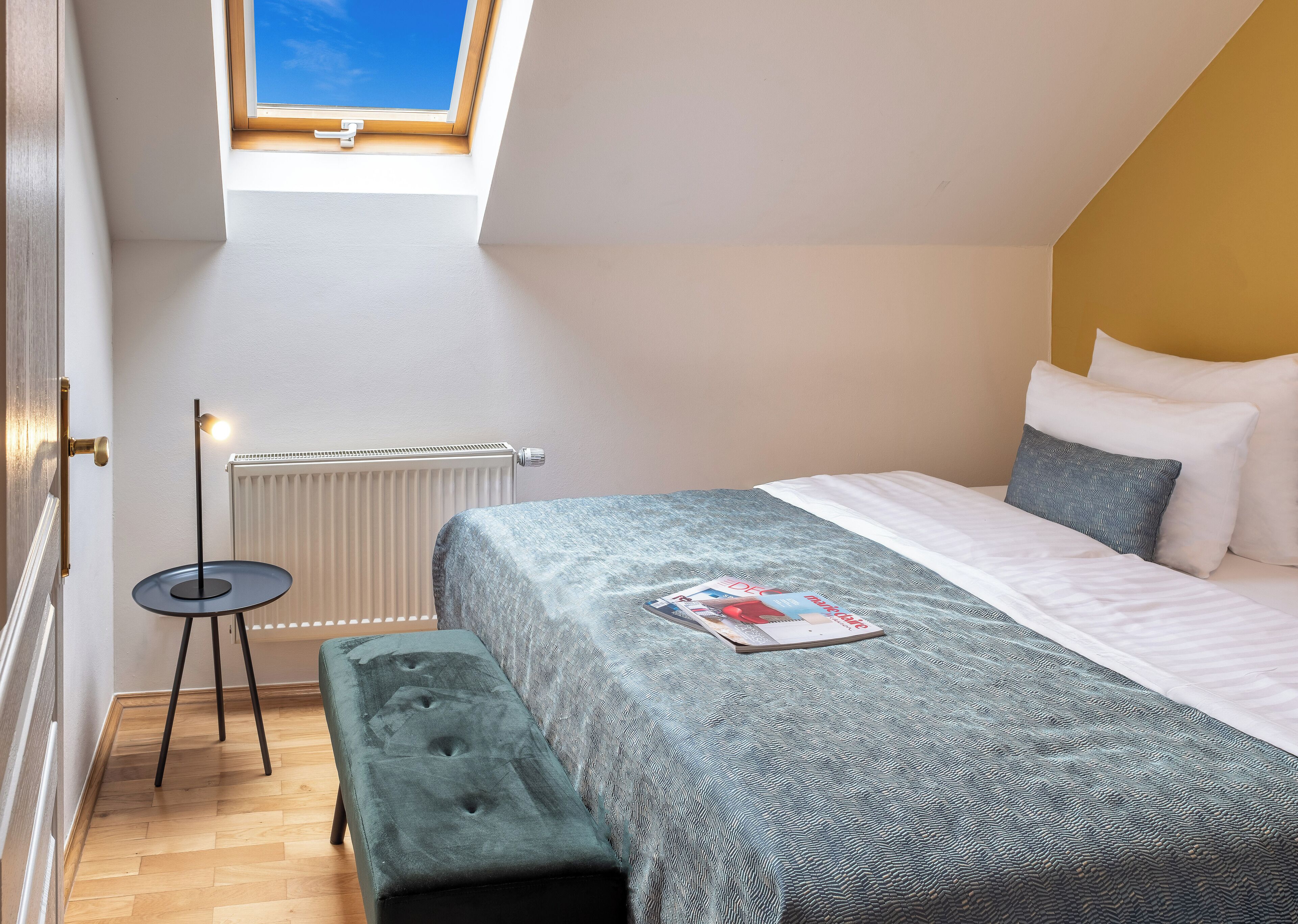 Une chambre avec un puits de lumière, un lit avec des oreillers, un banc et une lampe sur pied.