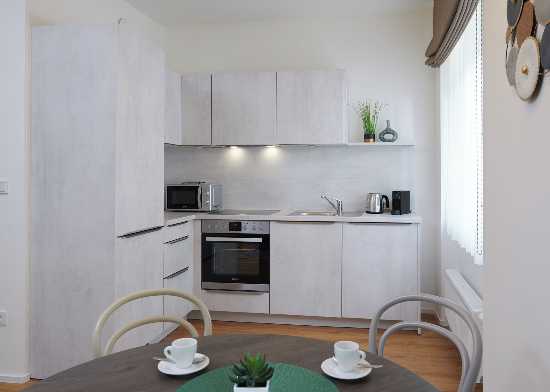 Une cuisine moderne avec des armoires blanches, un four encastré, un four à micro-ondes et une machine à café. On y trouve une table à manger avec deux chaises et une petite plante posée sur la table.