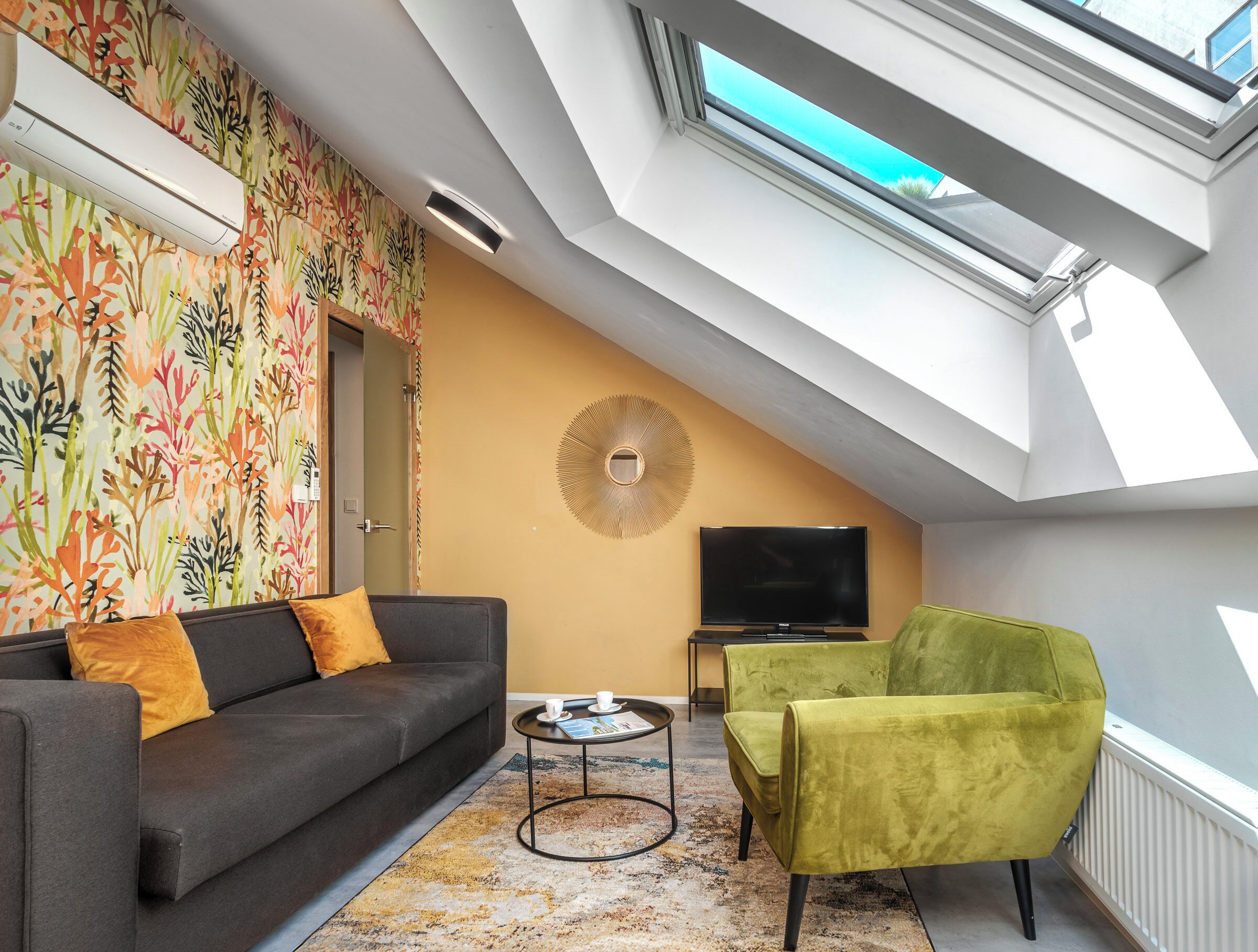 Ein modernes Wohnzimmer mit schrägverdeckter Decke, einem Flachbildfernseher, einem Sofa, einem grünen Sessel und einer Wand mit Muster.