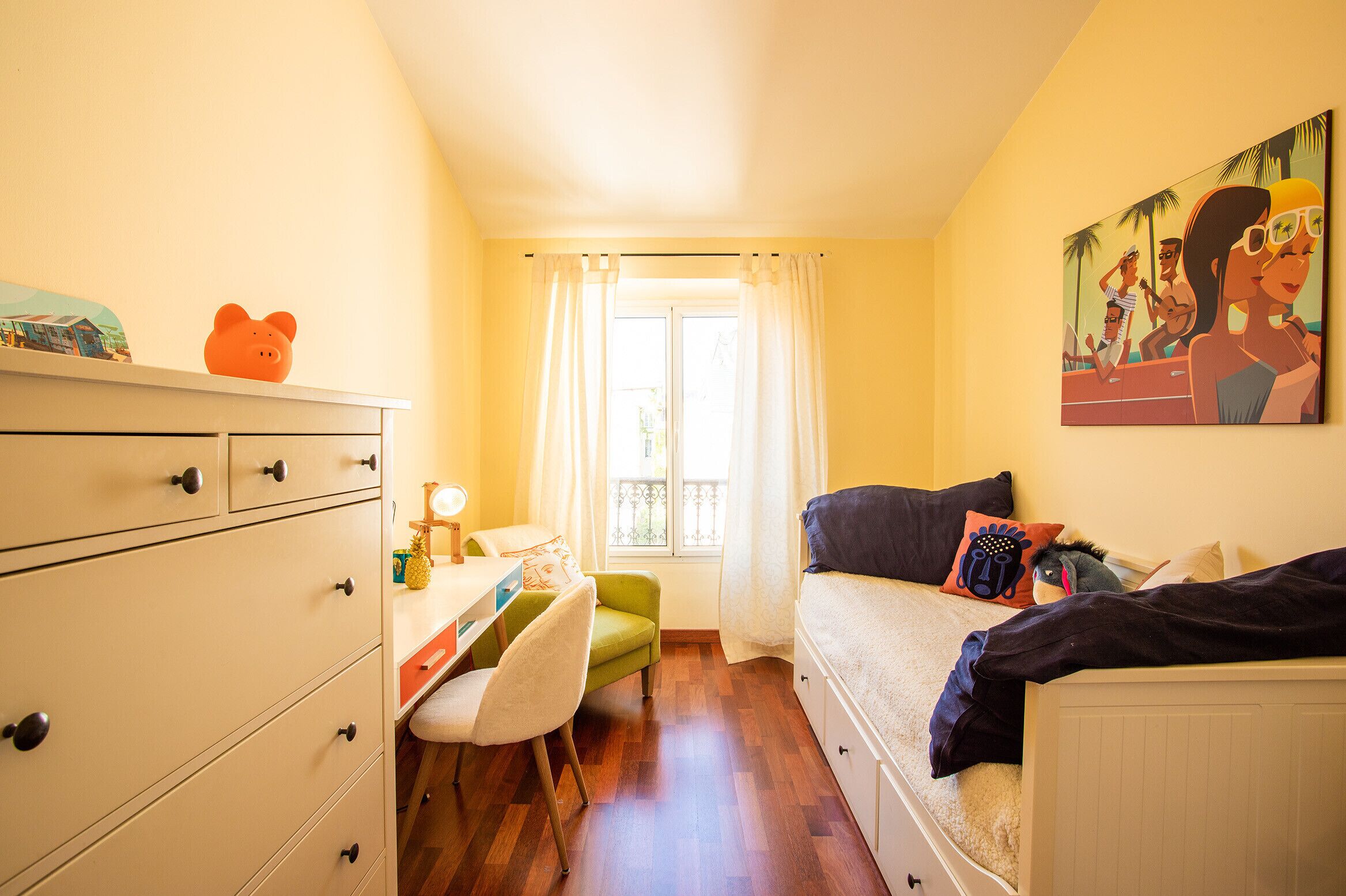 A small, cozy bedroom with a bed, a desk, and a chair. There is a window with curtains, a painting on the wall, and a dresser with drawers.