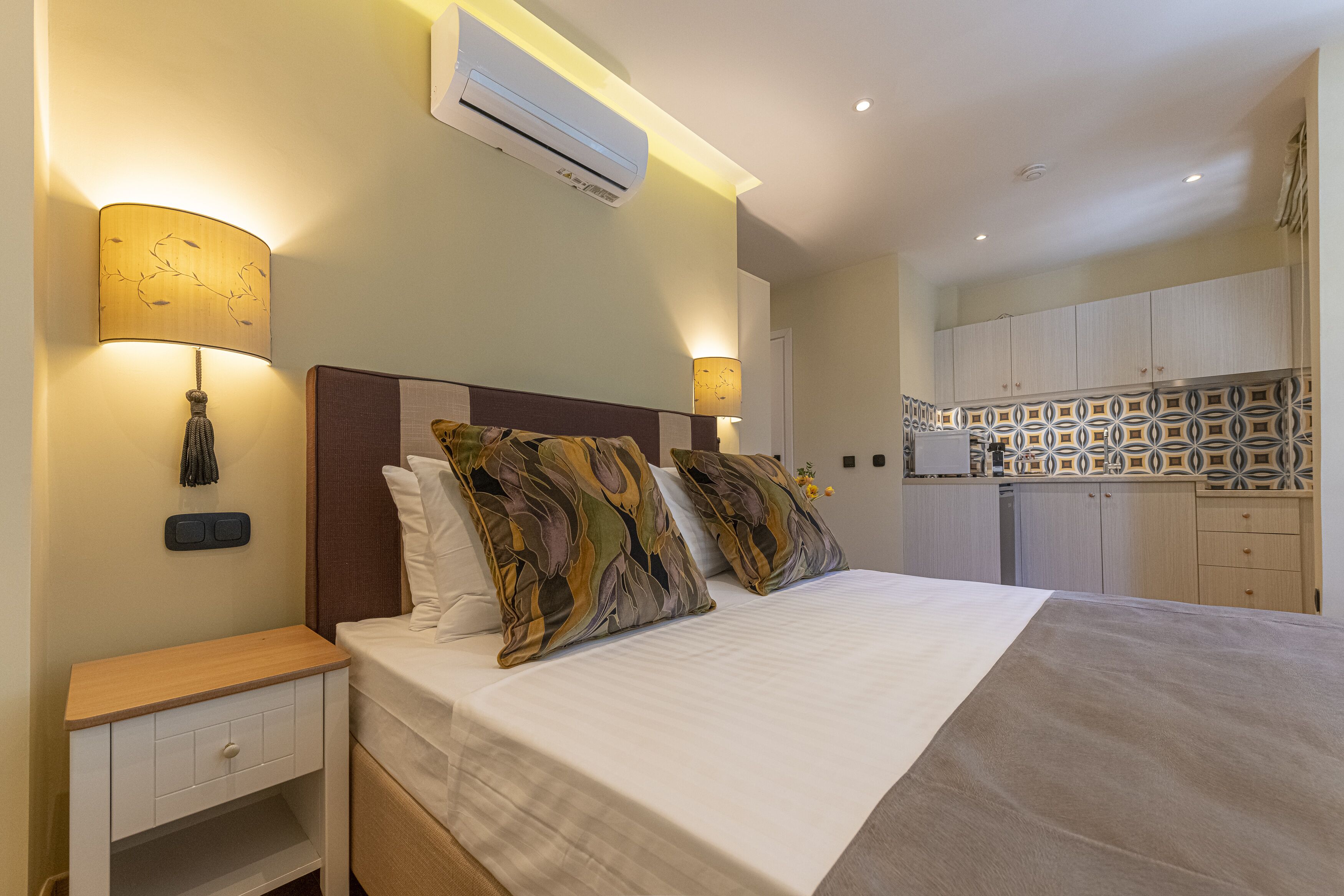 Une chambre d’hôtel avec un lit, des lampes de chevet, une table de nuit, un climatiseur et, au fond, un coin cuisine.