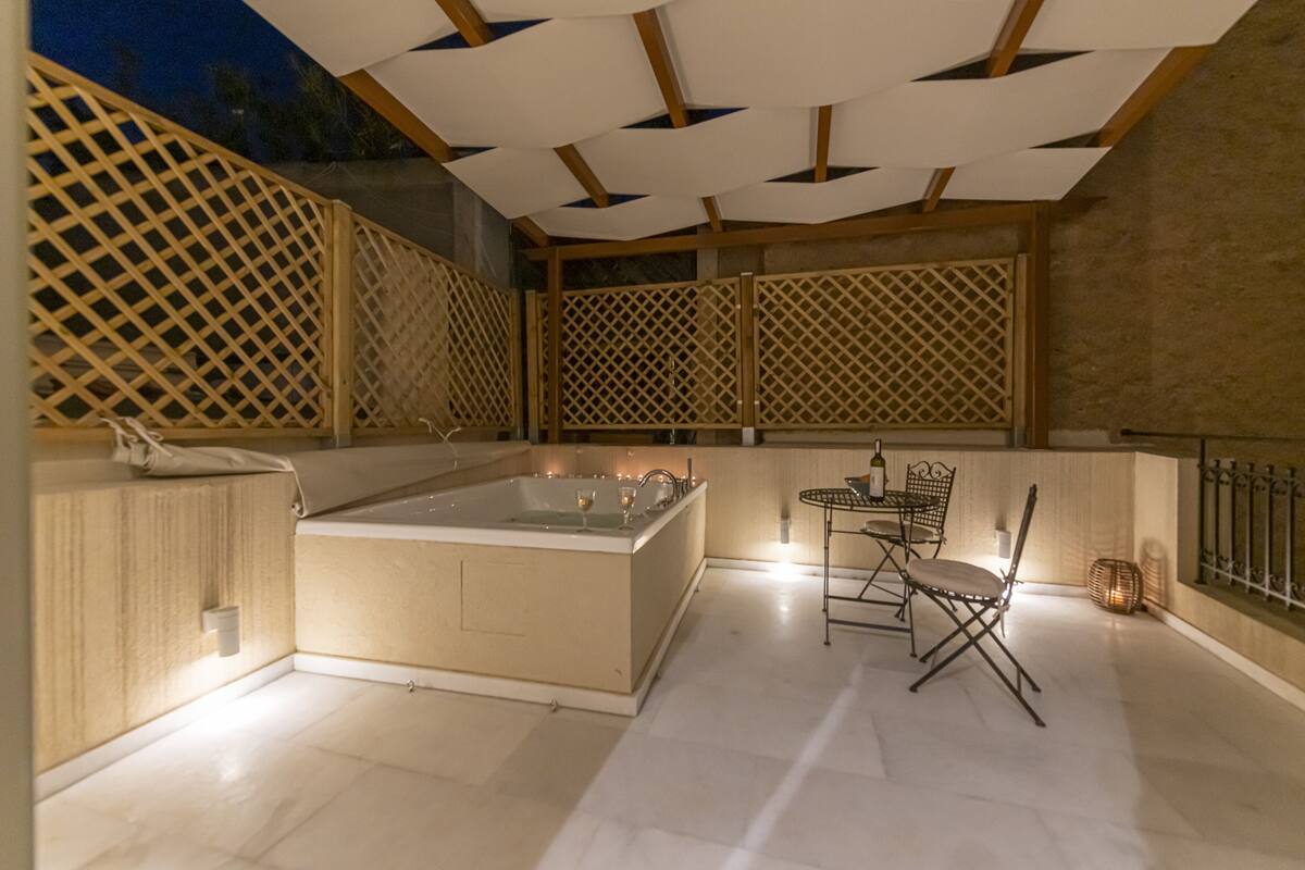 A modern outdoor bathroom with a freestanding bathtub, a small table with chairs, and a decorative lantern.