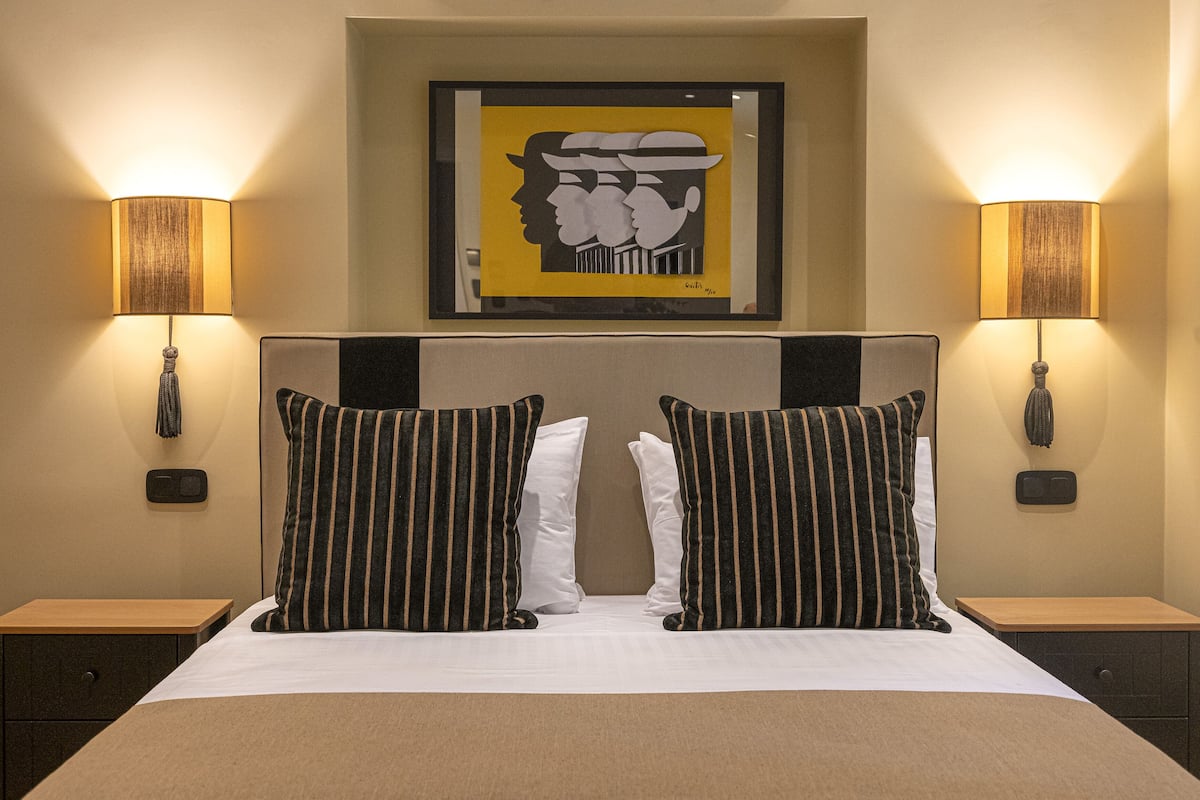 A hotel room with a large bed, two bedside lamps, a framed artwork above the bed, and a small nightstand with a phone and a switch.