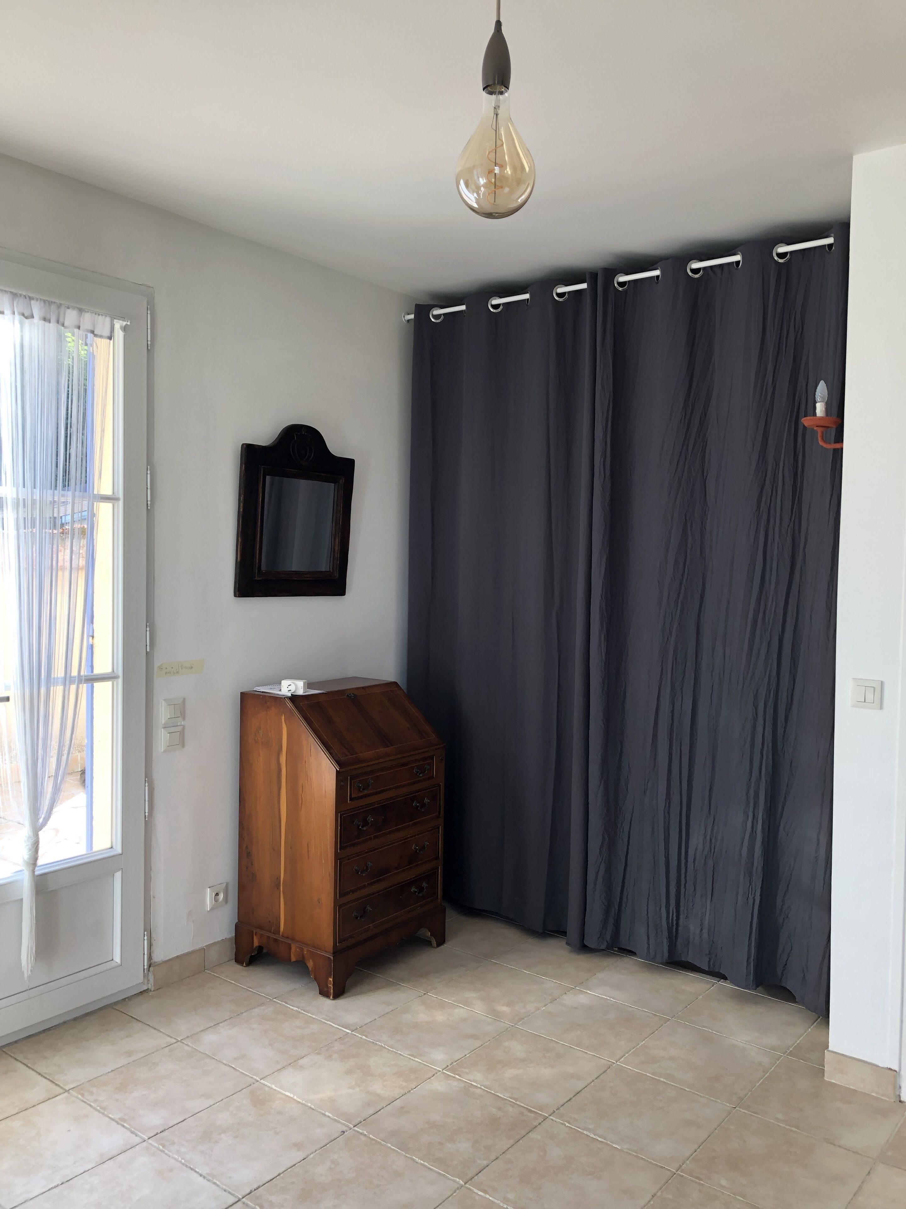A room with a wooden dresser, a dark curtain, a hanging light fixture, and a window with sheer curtains.
