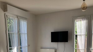 Une pièce avec une grande table en bois, un radiateur blanc, un téléviseur fixé au mur et une porte-fenêtre coulissante avec des rideaux.