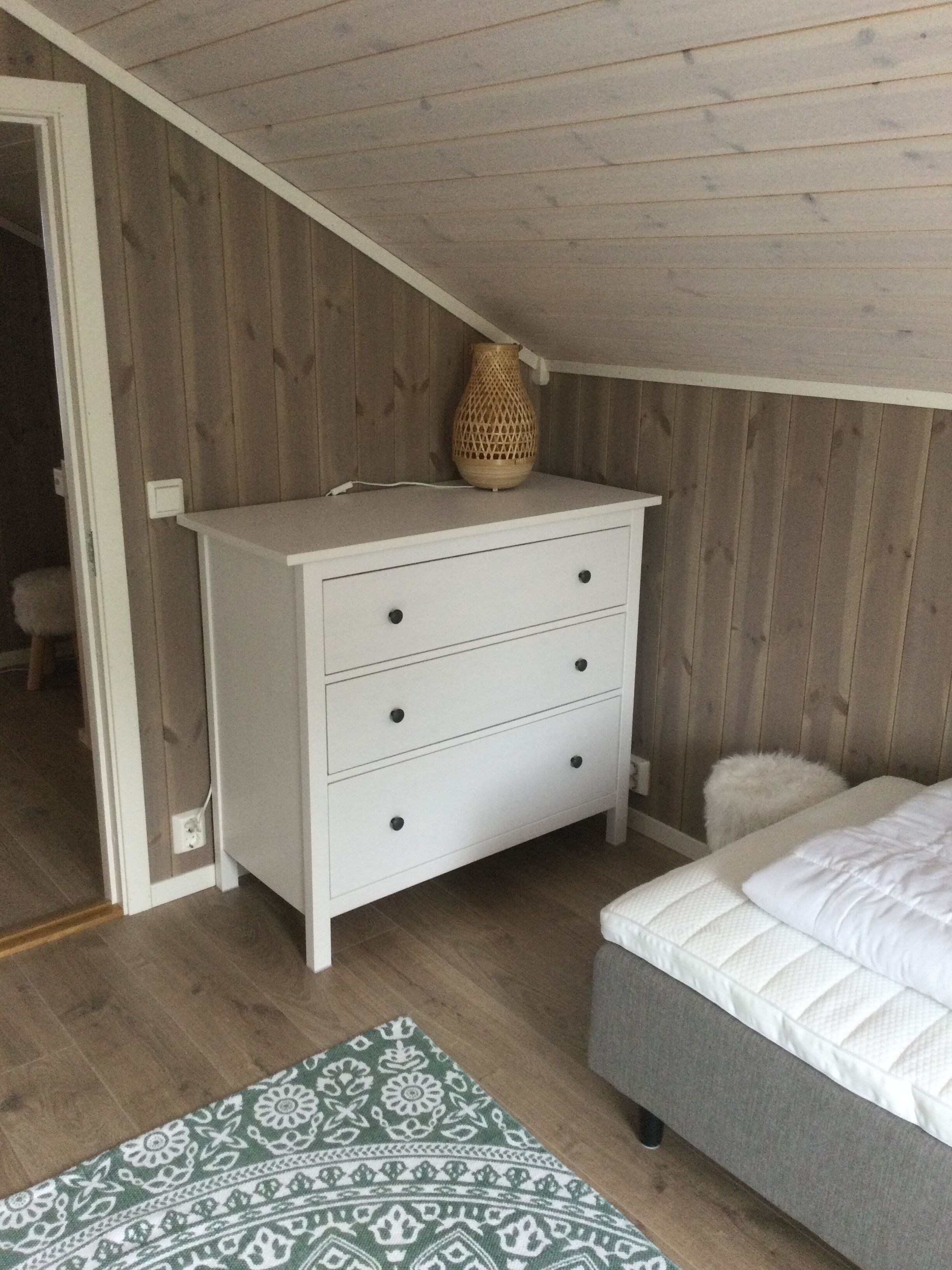 Un dormitorio con un tocador blanco, una alfombra estampada, una cama gris y un techo de madera.