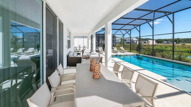 Un espace piscine extérieur moderne doté d’un patio fermé par des baies vitrées, de chaises longues et d’une table à manger.