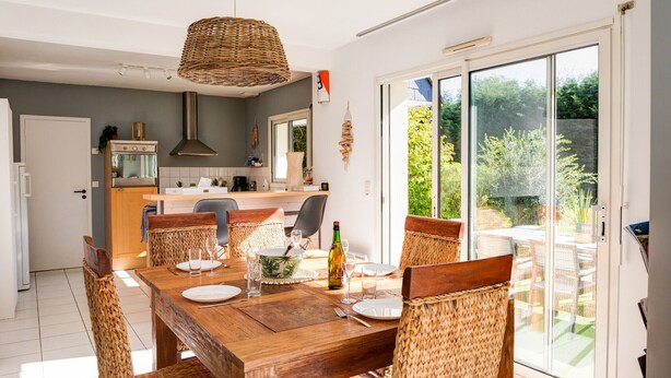 Zona pranzo con tavolo in legno e sedie in vimini, una cucina sullo sfondo e una porta scorrevole in vetro che dà su un giardino.