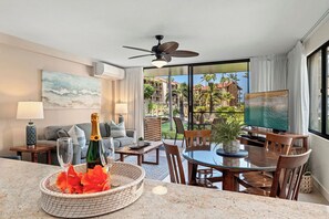 A hotel room with a balcony, a dining area, a sofa, a TV, and a ceiling fan. - Oceanview - Papakea H107 AC, King Bed, Ground Corner Unit (Lahaina)