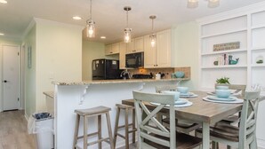 A kitchen with white cabinets, a black refrigerator, and a microwave. There are three hanging pendant lights, a wooden island with stools, and a dining table set with blue dishes.