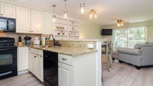 A kitchen with white cabinets, granite countertops, a black stove, and a built-in dishwasher. There is a dining area with a gray sofa and a TV mounted on the wall.