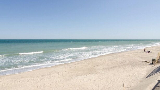 A beach with a clear sky, calm sea, and a few people and a lifeguard chair on the sand.