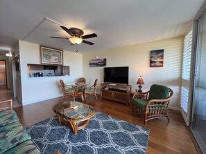A living room with a round glass coffee table, wicker chairs, a television, and a ceiling fan. - Kamaole Beach Royale #304, Panoramic Ocean View of Kam Beach #1, Full Kitchen (Kihei)
