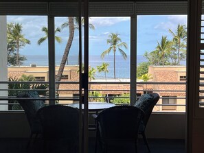 A room with a view of the ocean and palm trees through large windows. There are two wicker chairs and a small table. - Kamaole Beach Royale #304, Panoramic Ocean View of Kam Beach #1, Full Kitchen (Kihei)