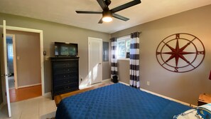 Un dormitorio con cama, cómoda, ventilador de techo, televisor, ventana con cortinas y un adorno en la pared.