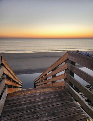Un paseo marítimo de madera que lleva a una playa con un mar tranquilo y una puesta de sol al fondo.
