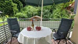 A round table with a white tablecloth, two chairs, a bottle, and two glasses on a balcony with a green umbrella.