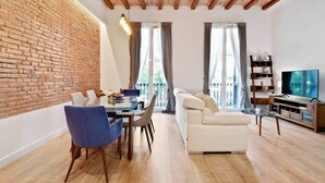 Un salon moderne avec un mur en briques, des poutres en bois au plafond, un canapé blanc, une table à manger avec des chaises et un téléviseur.