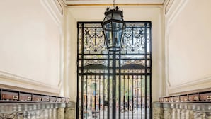 Un vaste hall avec un sol en marbre, une grande porte richement décorée et surmontée d’une grille métallique, et une lanterne suspendue.