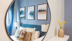 Une chambre à coucher avec un lit, des oreillers, une table de chevet avec une lampe et un vase de fleurs. Trois œuvres d’art encadrées ornent le mur.