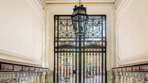 Un vaste hall avec un sol en marbre, une grande porte richement décorée et surmontée d’une grille métallique, et une lanterne suspendue.