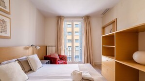 A hotel room with a bed, a red armchair, a wooden wardrobe, and a window with curtains. - Paseo de Gracia | Classy 3bed 3bath apartment (Barcelona)