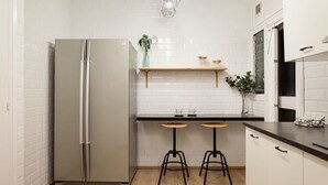 A compact kitchen with a stainless steel refrigerator, a white tiled backsplash, a wooden countertop, and two stools.
