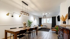 A modern living room with a wooden dining table, black chairs, and a sofa. There is a TV on the wall and a plant on the floor.