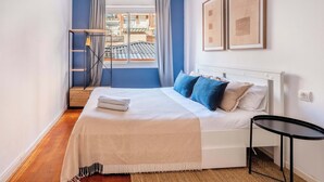 Une chambre à coucher avec un lit, une petite table, une fenêtre avec des rideaux et des œuvres d’art accrochées au mur.
