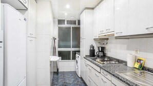 Une cuisine compacte avec des meubles blancs, un lave-linge encastré, un plan de travail en granit et une fenêtre avec des stores.