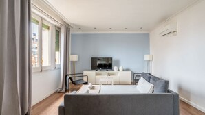 Une chambre d’hôtel moderne avec un canapé gris, un petit bureau, un téléviseur à écran plat et une fenêtre avec des rideaux.