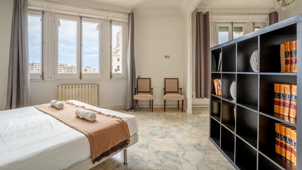 Une chambre d’hôtel avec un lit, deux chaises, une bibliothèque et une vue sur les immeubles à travers de grandes fenêtres.