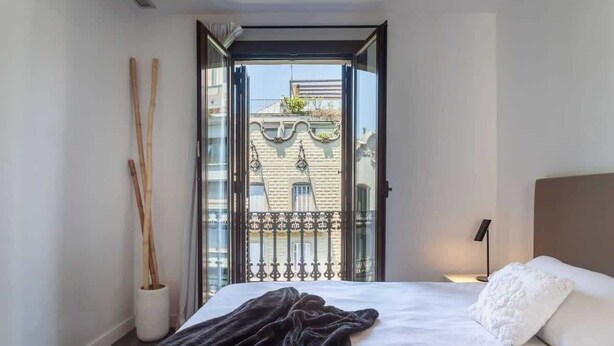 A bedroom with a bed, a nightstand, a lamp, and a view of a building through an open door. - 3-bedroom penthouse with terrace in Eixample (Barcelona)
