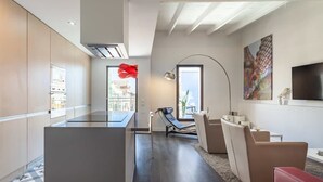 A modern kitchen with a central island, a dining area with a red chair, and a living area with a sofa and a TV. - 3-bedroom penthouse with terrace in Eixample (Barcelona)