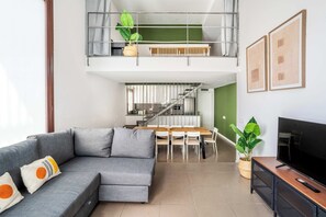 Een moderne woonkamer met een grijze modulaire bank, een eettafel met stoelen, een tv op een houten dressoir en een trap naar een hoger niveau.
