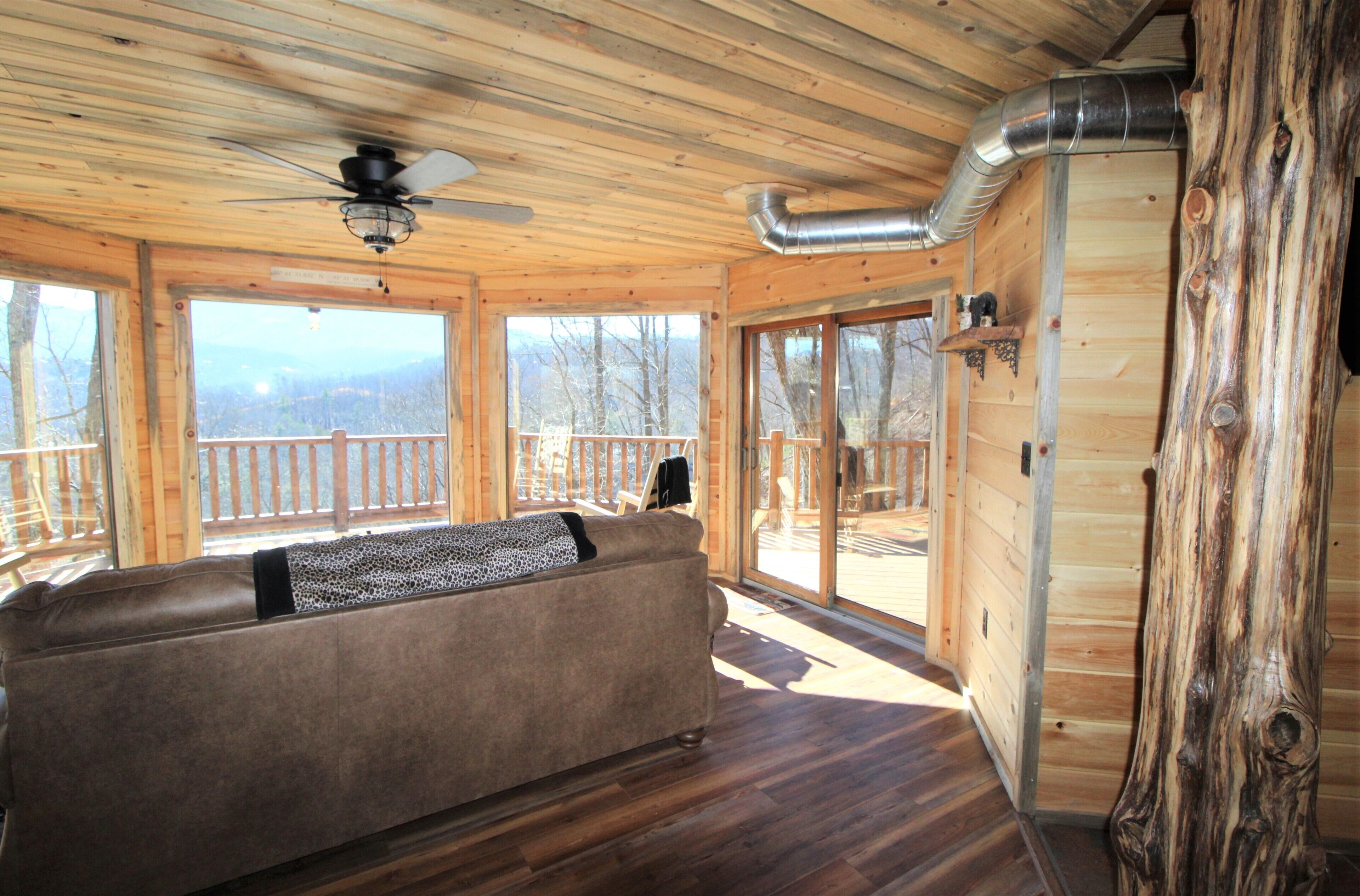 A wooden cabin interior with a sofa, large windows, and a ceiling fan.
