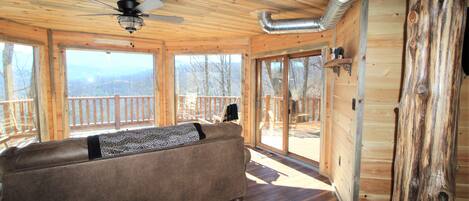 A wooden cabin interior with a sofa, large windows, and a ceiling fan.