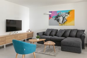 A modern living room with a grey sectional sofa, a blue armchair, a wooden coffee table, a flat-screen TV mounted on a white wall, and a colorful abstract painting above the sofa. - Villa Brela Apartment 1 (Brela)
