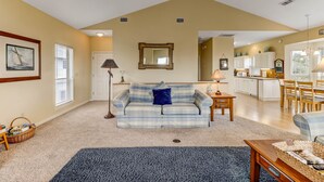 A living room with a sofa, a coffee table, and a rug. There is a kitchen in the background with white cabinets and a dining area with chairs.