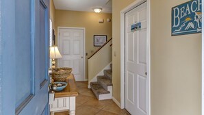 A hallway with a blue door, a white bench with a basket, a framed picture, a staircase, and a 'Beach' sign on the wall.