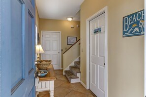 Um corredor com uma porta azul, um banco branco com cestas, um quadro emoldurado, uma escada e uma placa com a palavra 'Beach' na parede.