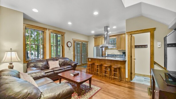 A spacious living room with a leather sofa, a coffee table, and a dining area with bar stools. A kitchen with wooden cabinets and a stainless steel range hood is visible. - Ridge Street Chalet B 2 Bedroom Townhouse by RedAwning (Breckenridge)